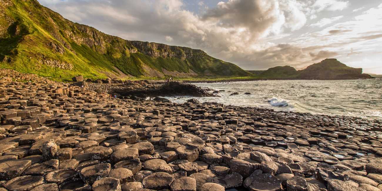 A wide landscape view of the interlocking geometric basalt columns at the Giant's Causeway with dramatic clouds over the green Antrim Coast cliffs